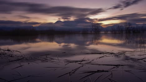 Dark-to-dawn-time-lapse-of-a-lake-with-snow-capped-mountains-in-the-background-and-the-sky-reflecting-off-the-lake---zoom-out-reveal