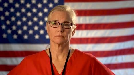 Medium-tight-portrait-of-a-healthcare-nurse-looking-concerned-and-sympathetic-walking-towards-the-camera-with-an-out-of-focus-American-flag