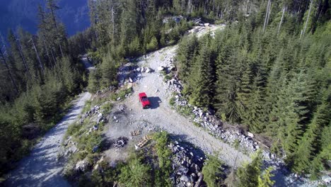 a-reverse-revealing-aerial-view-of-a-jeep-parked-on-a-backroad-trail-surrounded-by-trees-with-mountains-and-blue-sky-in-the-background