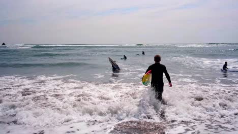 Joven-Corriendo-Hacia-El-Mar,-Motivado-Para-Surfear,-Mientras-Los-Niños-Toman-Clases-De-Surf