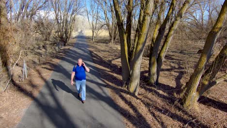 Older-man-on-a-nature-walk-can't-get-away-from-his-smartphone-and-talks-while-walking---slow-motion-aerial-view
