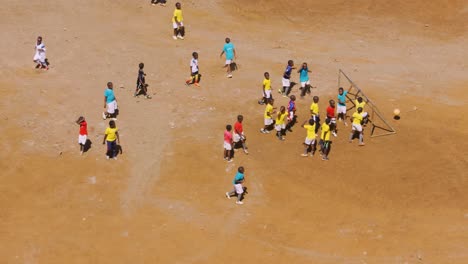 Group-of-young-black-african-children-playing-soccer-on-red-dirt-pitch-with-brightly-coloured-tops
