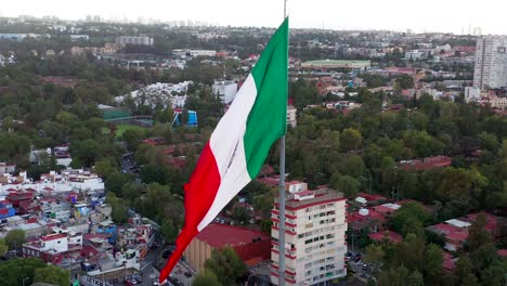 Circle-flight-by-large-majestic-patriotic-red,-white-and-green-Mexican-flag-waving-in-wind-above-downtown-Mexico-city-center,-buildings-and-San-Jeronimo-community,-on-cloudy-sky-day,-aerial