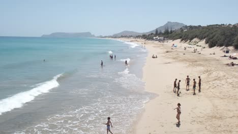 Summer-scene-of-people-swimming,-relaxing-and-kids-playing-soccer-by-ocean-sea-waves-on-Porto-Santo-island-sandy-beach-with-mountain-range-in-background,-Portugal,-handheld-pan-left