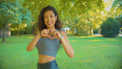 Young-Attractive-Middle-Eastern-Woman-Kissing-Camera-and-Showing-Heart-Hand-Sign-in-Exterior-of-Public-Park,-Slow-Motion