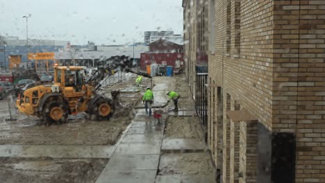 Wet-window-frame-with-construction-workers-operating-heavy-machinery-in-laying-a-sidewalk-in-front-of-newly-build-houses-on-a-rainy-day