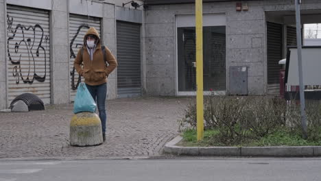 A-Man-In-Mask-And-Hoodie-Jacket-Waiting-To-Cross-The-Street-In-Arcore,-Italy,-wide-shot