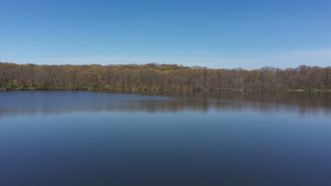 An-aerial-view-of-a-blue,-reflective-lake-with-bare-trees-lining-the-lake's-edge