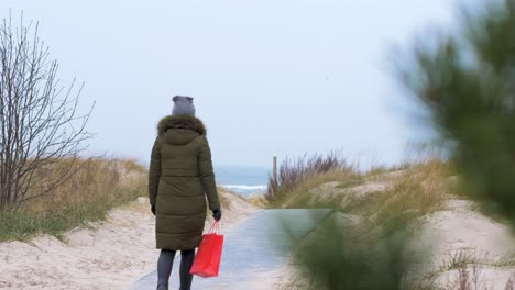 Young-woman-walking-along-the-beach-in-winter-overcast-day,-red-bag-in-right-hand,-out-of-focus-pine-tree-in-foreground,-handheld-medium-shot