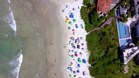 Luftaufnahme-Von-Oben-Nach-Unten-Auf-Den-Strand-Von-Bombinhas-Mit-Menschen-Und-Sonnenschirmen-Und-Einem-Hügel-Auf-Der-Rechten-Seite