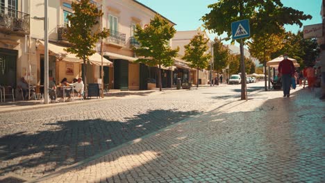 Portugal-Algarve-Loule-Calle-De-Piedra-Tradicional-Con-Peatones-Caminando,-Terrazas,-Autos-Pasando-Por-El-Sol-De-La-Mañana-4k