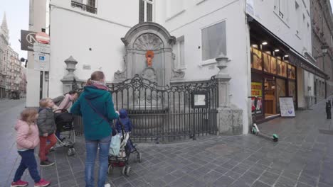 Family-Wearing-Face-Mask-Visit-Manneken-Pis-During-COVID-19-In-Central-Brussels,-Belgium---Mothers-Pushing-Baby-Stroller-Towards-Bronze-Fountain-Sculpture