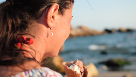 Closeup-slowmo-shot-of-a-beautiful-woman-with-a-natural-look-licking-delicious-ice-cream-on-the-beach-while-the-waves-are-breaking-around-and-the-sun-is-illuminating-her-face
