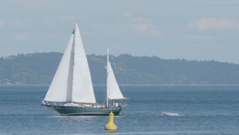 A-green-two-masted-sailboat-pulls-a-dingy-through-Commencement-Bay,-Puget-Sound,-and-passes-a-yellow-buoy