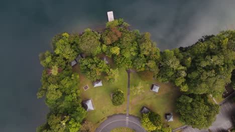 Moving-drone-video-looking-down-on-a-lush-park-area-next-to-a-volcanic-crater-lake-with-clouds-reflecting-on-the-calm-water