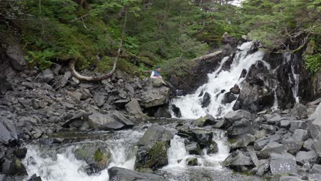 Backpacker-Traveler-Sits-In-Front-Of-Cascading-Water-Among-Rocks-Covered-With-Moss-At-Forest-Park-In-Alaska