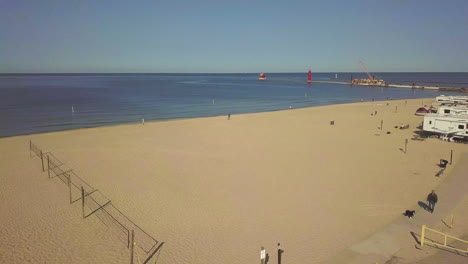 Moving-drone-shot-past-American-flag-and-beach-volleyball-courts-over-campground-located-next-to-large-sandy-beach-with-a-large-lake-in-the-background-with-a-pier-under-construction-with-a-lighthouse