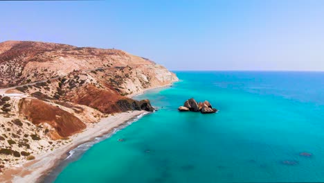 Aerial-view-of-Aphrodite's-Rock-as-camera-pulls-away-along-the-beach-and-coastline-with-beautiful-vivid-blue-sea