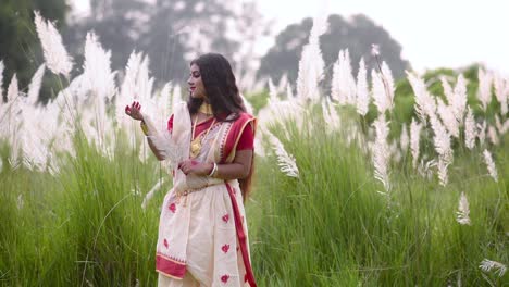 A-calm-and-beautiful-Indian-woman-is-happy-and-plays-with-white-grass-called-saccharum-spontaneum-in-a-field,-gold-jewellery,-slow-motion