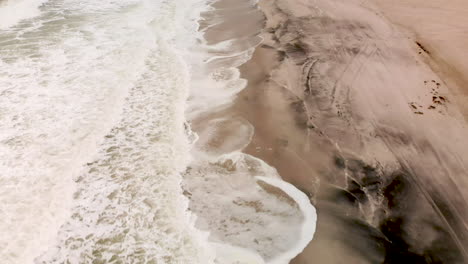Aerial-shot-of-flying-backwards-looking-down-at-waves-breaking-onto-a-sandy-beach