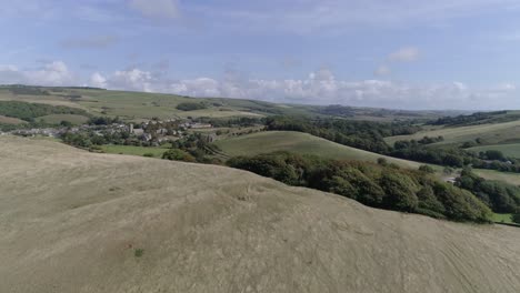 Low-tracking-aerial-moves-forward-fast-up-above-a-hill-to-reveal-the-village-of-Abbotsbury,-in-the-Dorset-countryside