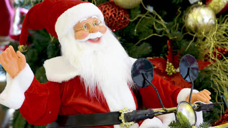 Muñeco-De-Santa-Claus-Saludando-Y-Montando-Una-Moto-Frente-Al-árbol-De-Navidad