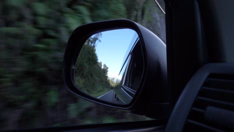 Slowmo---Car-driving-down-the-West-Coast,-New-Zealand-rear-view-mirror-with-mountains,-trees-and-blue-sky