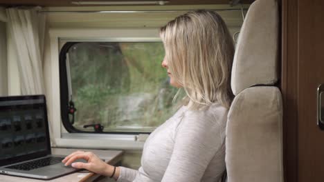 Beautiful-blond-girl-sitting-in-campervan-at-table-working-on-laptop-with-trees-and-nature-in-background