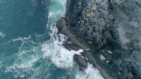 Top-down-aerial-of-a-craggy-dark-coloured-rocky-cliff-with-powerful-waves-crashing-creating-foam-from-the-dark-blue-Atlantic-water
