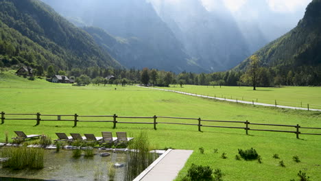 Panoramic-view-in-Logarska-valley,-Slovenia,-green-meadows-with-forest-and-high-mountains-in-background,-natural-swimming-pool-with-sunchairs-in-front