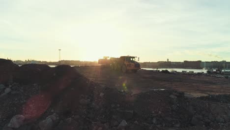 Static-aerial-shot-of-an-excavator-digging-and-loading-dirt-into-a-truck-with-sunset-behind