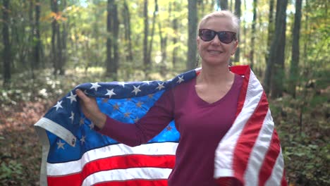 Pretty,-blonde-woman-walking-through-a-sunlit-forest-holding-out-a-flag-and-then-wrapping-it-around-her-as-she-smiles-and-looks-at-the-camera