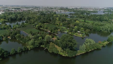 Aerial-Slomo-Wide-shot-of-Dutch-Countryside-surrounded-with-Small-Rivers,-Green-Trees-and-Bushes,-tilting-up,-Revealing-Small-Town-in-background