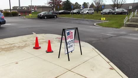 Vote-here-sign-with-American-flag-blowing-in-the-wind,-wide