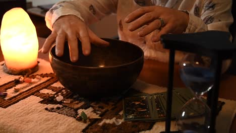hourglass-marking-the-time-with-its-blue-sand,-with-a-man-moving-his-hands-above-a-tibetan-bowl-near-tarot-cards-near-some-stones-on-a-carpet,-near-a-salt-lamp