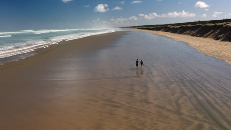 Aerial-view-of-a-young-couple-walk-hand-in-hand-along-the-ninety-mile-beach,-New-Zealand
