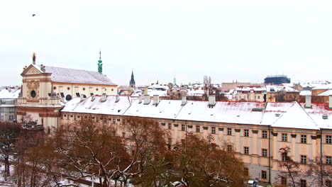 Prague-winter-flight-snow-over-trees-and-houses-birds-flying-aerial-drone-next-to-church