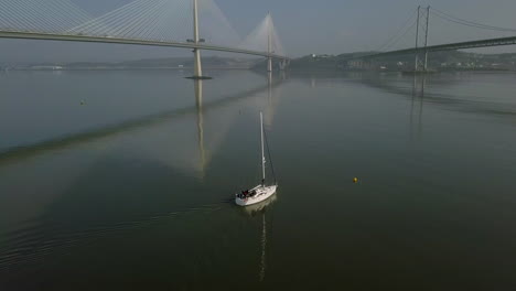 Aerial-footage-of-a-sailing-boat-travelling-between-the-Forth-bridges-at-South-Queensferry-on-a-sunny-day-in-West-Lothian,-Scotland