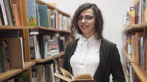 Friendly-teacher-holds-a-book-in-the-library-and-smiles-at-the-camera
