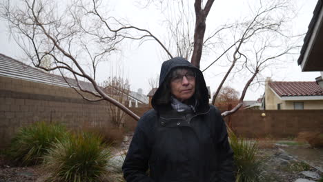 A-beautiful-old-woman-wearing-a-black-raincoat-and-hood-during-a-winter-rain-storm-as-drops-fall-from-the-cloudy-sky-in-slow-motion