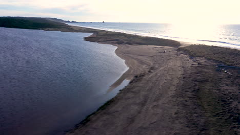 Drone-flying-over-the-edge-of-Floras-Lake,-Oregon,-showing-the-Pacific-Ocean-divided-by-only-a-thin-strip-of-land,-as-well-as-Blacklock-Point-in-the-background