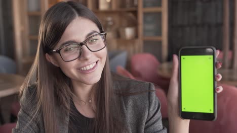 Adorable-young-business-lady-in-a-glasses-is-sitting-in-a-cafe,-holding-a-phone,-smiling-and-demonstrated-a-green-screen-of-the-phone
