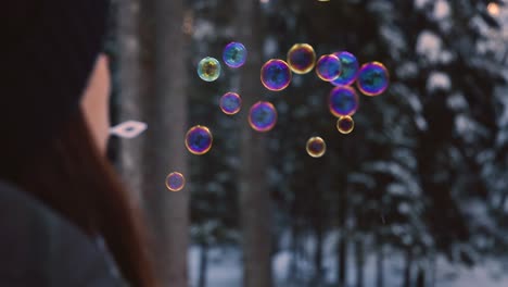brunette-woman-with-winter-clothes-blowing-rainbow-colored-soap-bubbles-flying-suspended-in-the-air,-with-a-snowy-forest-in-the-background
