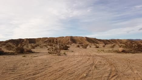Panning-view-of-sandy-surface-marked-with-off-road-vehicle-tire-tracks-and-rising-hills-beyond