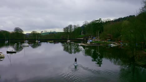 Beautiful-aerial-view,-footage-of-middle-aged-man-paddle-boarding-on-Rudyard-Lake-in-the-Derbyshire-Peak-District-National-Park,-popular-holiday,-tourist-location-with-peaceful-calm-water
