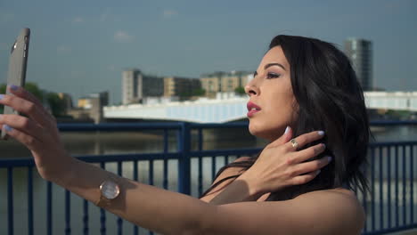 Schöne-Brünette-Hispanische-Modelfrau-Schaut-Auf-Ihr-Telefon-Und-Macht-Selfies-Neben-Der-Themse-In-London-Bei-Hellem-Tageslicht-Des-Sommers,-Im-Vereinigten-Königreich