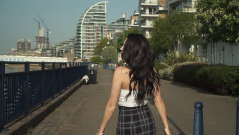 Attractive-and-playful-latina-woman-with-black-wavy-hair-walking-on-the-banks-of-the-Thames-river-in-London,-looking-at-the-camera,-doing-follow-me-gesture,-happy-with-a-beautiful-smile