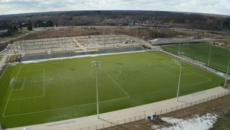 Drone-view-of-empty-soccer-field-during-winter-as-the-snow-is-thawing-with-parking-lot-and-road-in-the-distance