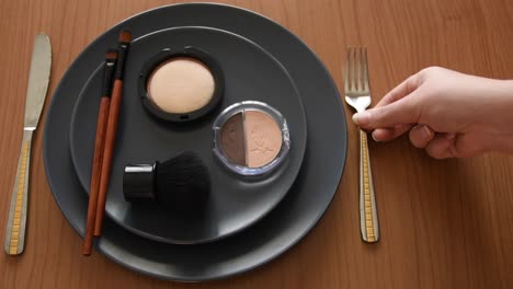 minimal-concept-background-with-a-white-female-hand-putting-a-fork-on-one-side-of-a-grey-plate-with-make-up,-brushes-and-powders-on-it,-on-the-other-side-there-is-a-knife,-on-a-wooden-table