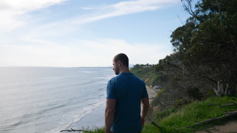 A-fit-young-man-walking-along-the-edge-of-an-ocean-cliff-looking-down-on-the-beach-on-a-sunny-day-in-nature-during-a-hike-in-Santa-Barbara,-California-SLOW-MOTION